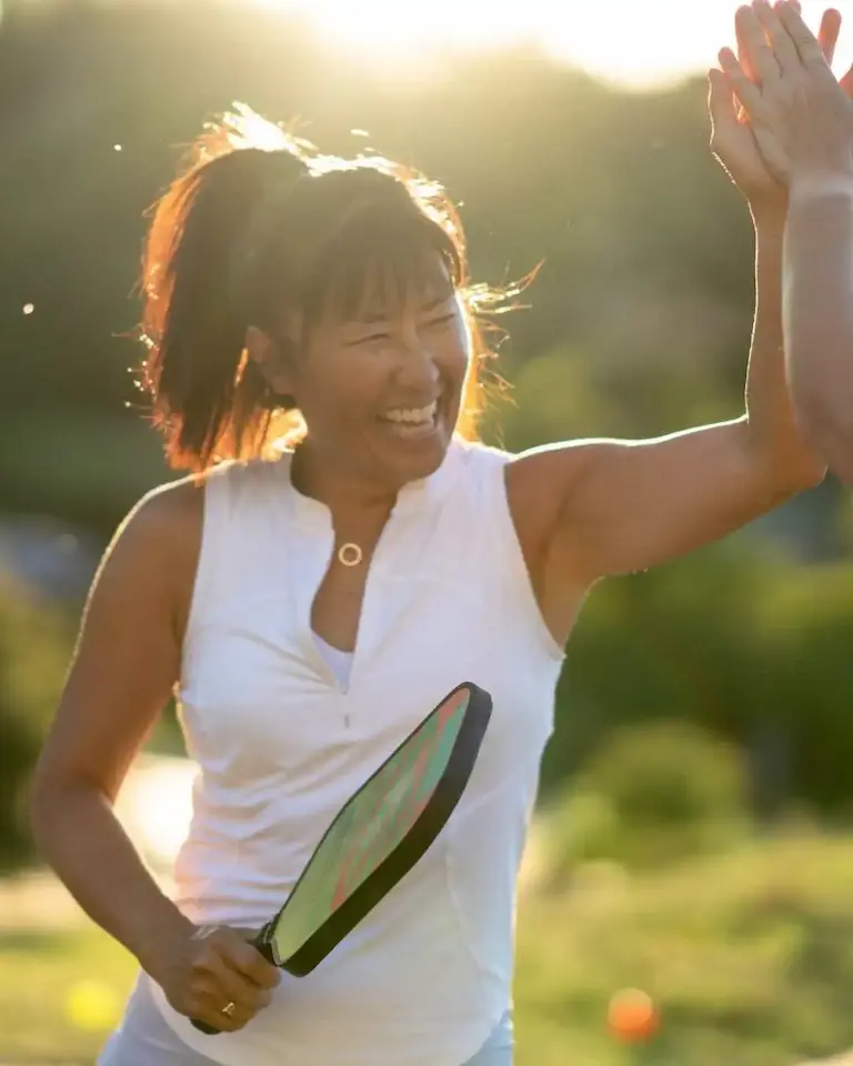 Home 1 Femme souriante jouant au pickleball en extérieur, raquette à la main, donnant un high five à son partenaire – scène typique d'un match convivial
