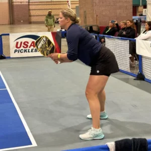 Clijsters Kim Athlète de profil, en position d’attente avec une pagaie de pickleball, sur un court intérieur ; panneau « USA Pickleball » en arrière-plan.