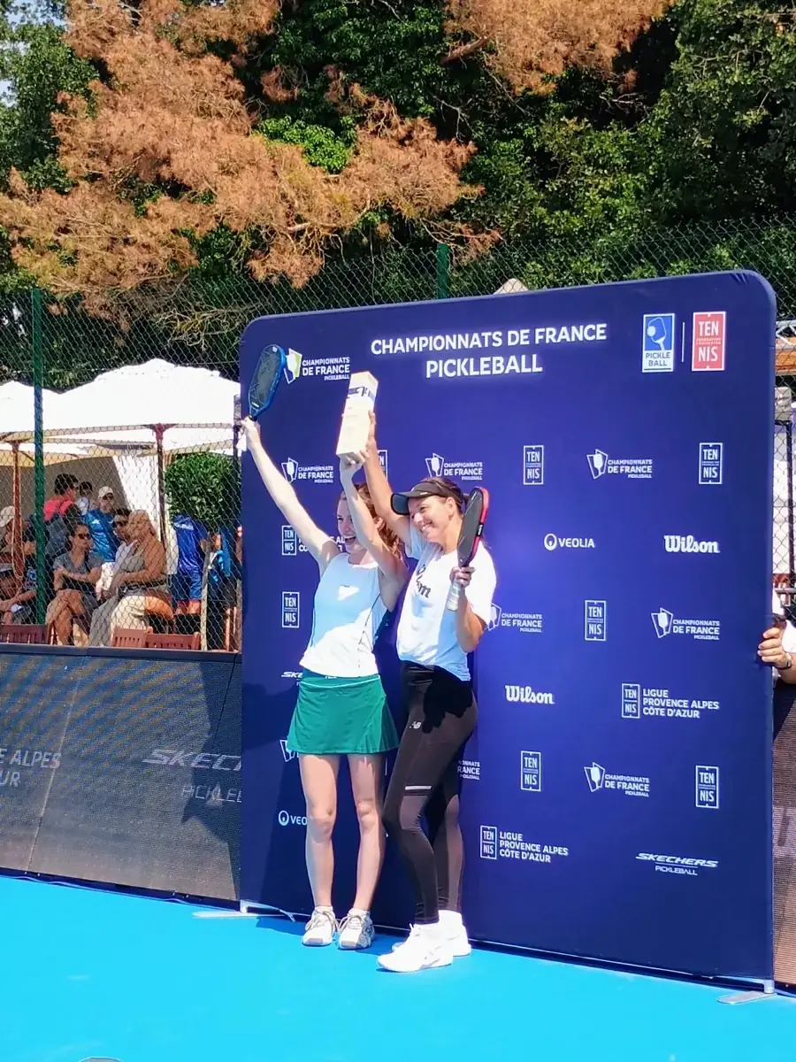 Deux vainqueurs lèvent leur trophée devant le mur officiel des Championnats de France de pickleball 2026